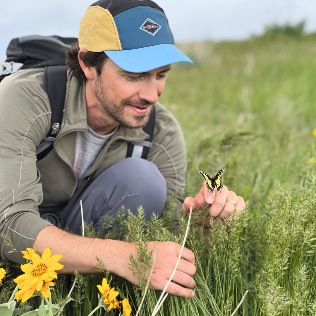 Man crouches in a field near flowers while looking at a butterfly that's landed on his hand.
