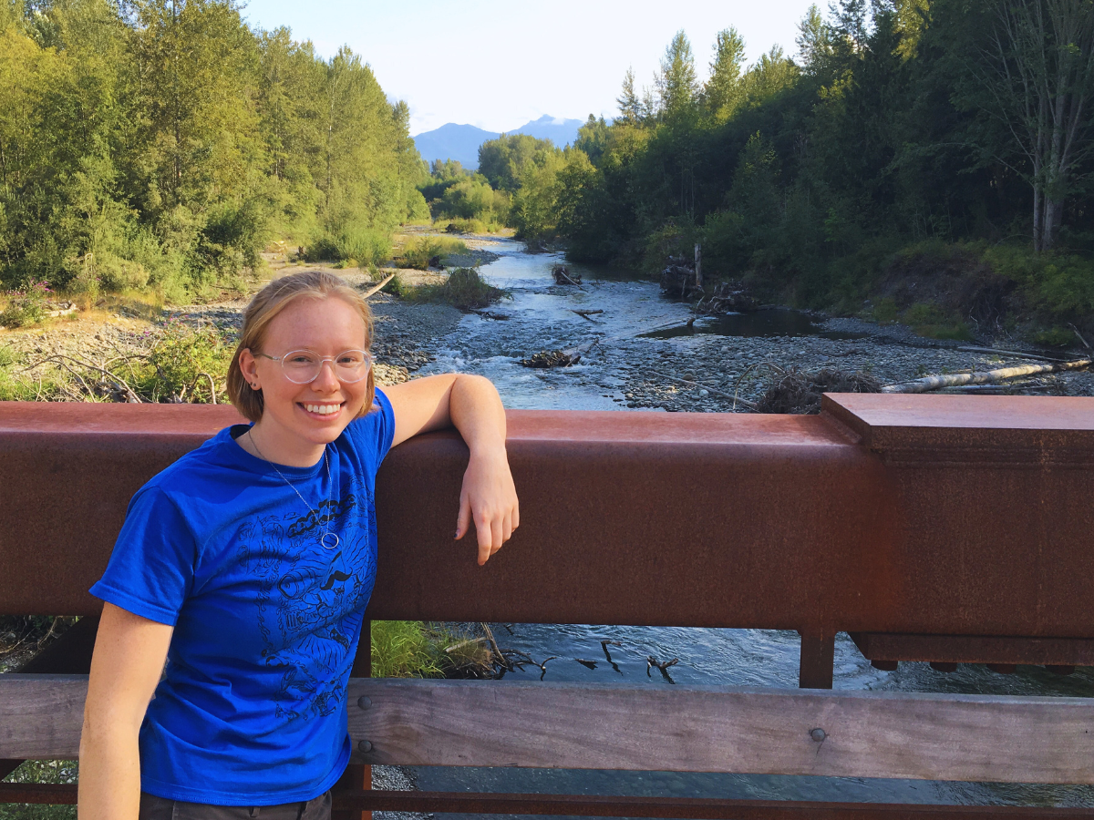 Woman stands on bridge over a flowing river with wooded banks and mountains in the distance.