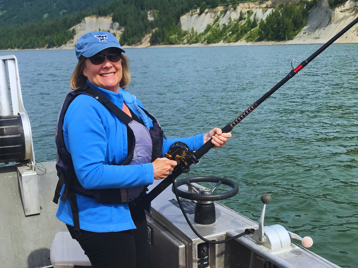 Lorri Bodi smiles and holds a fishing rod on a boat with water and forested hills in the background.