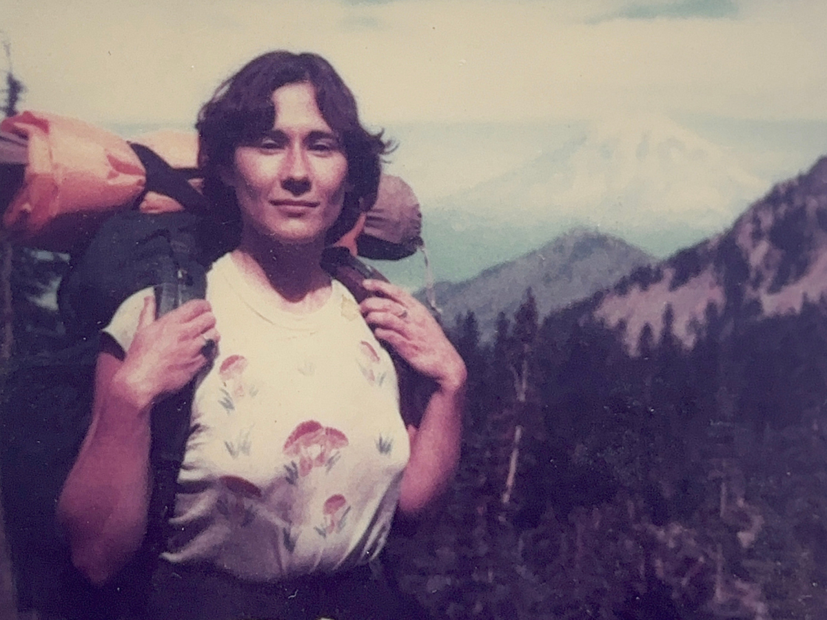 A faded photo of Lorri Bodi smiling while backpacking through a mountain forest in the 1980's.