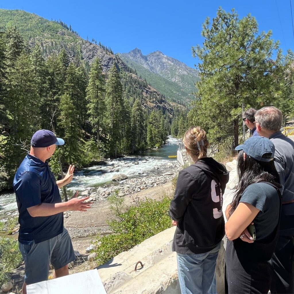 A man gestures towards a river flowing through trees behind him as a group listens and looks at the river.