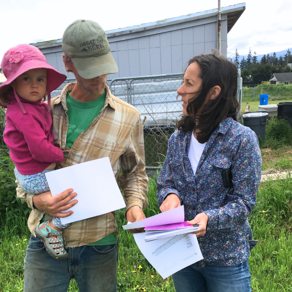 Two people talk while pointing at paperwork they're holding in a yard; a baby sits in the taller person's arm.