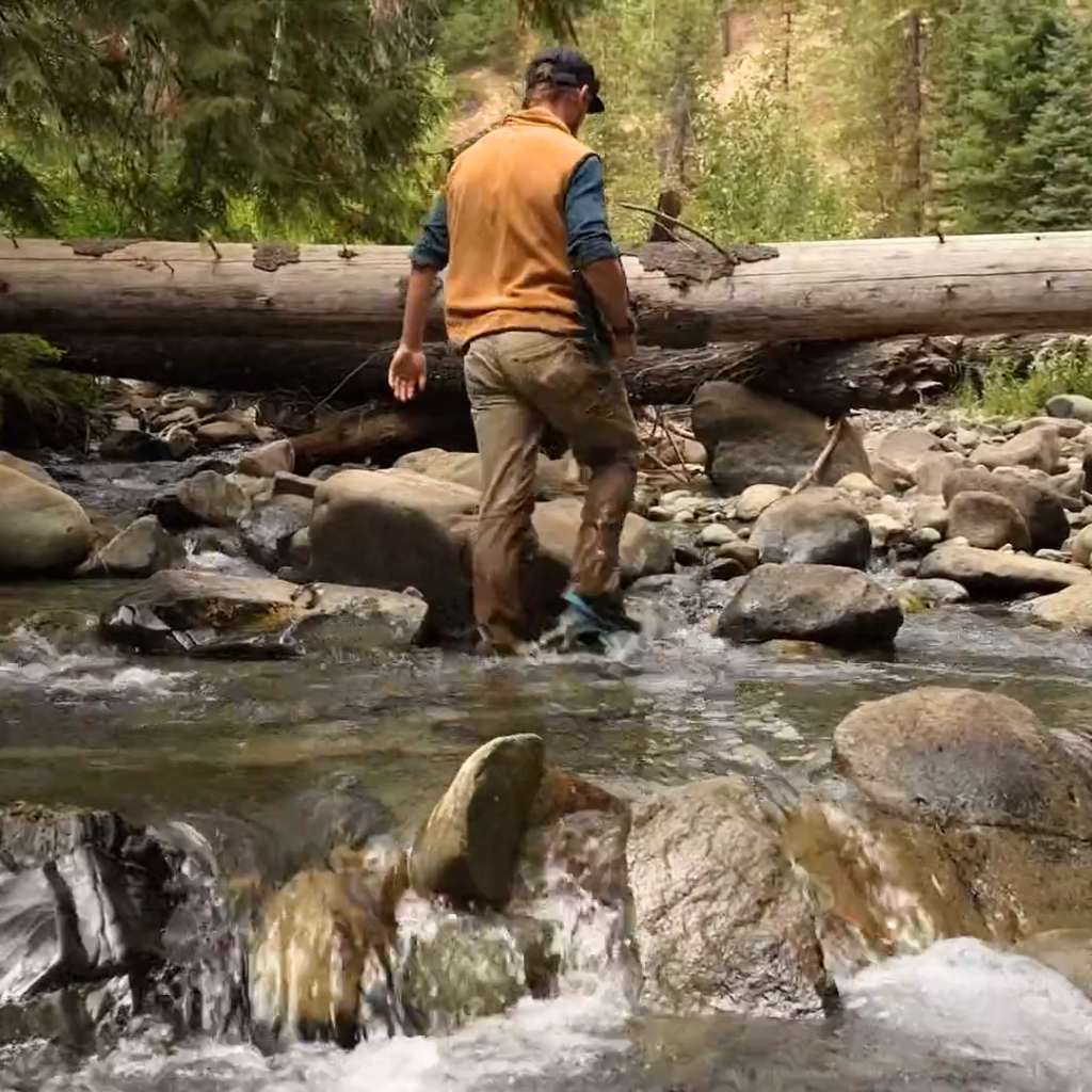 A man wades through a flowing river in the woods, walking away from the camera.