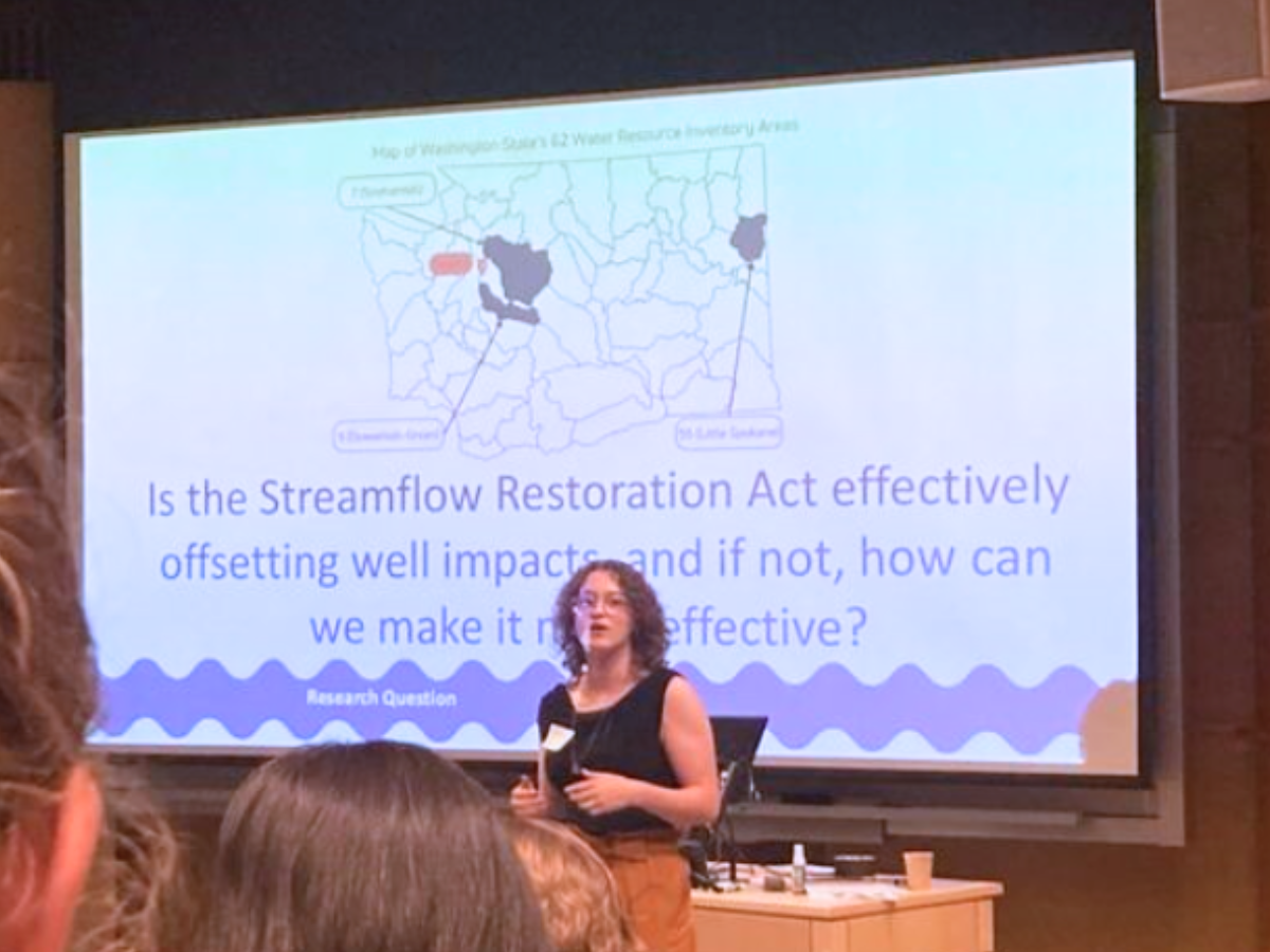 Woman presents in front of a presentation slide that says, "Is the Streamflow Restoration Act effectively offsetting well impacts and, if not, how can we make it effective?"